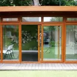 Zadaszona konstrukcja z dużymi, przesuwnymi oknami w drewnianych ramach, widok na trawnik i ogród z krzesłami wewnątrz.