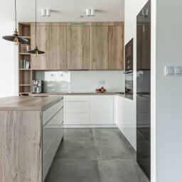 byMGdesigns - Nowoczesna kuchnia z wyspą, drewniane szafki górne i białe dolne, szare płytki podłogowe, lampy wiszące z żarówkami Edisona, ekspres do kawy na blacie, minimalistyczny design.