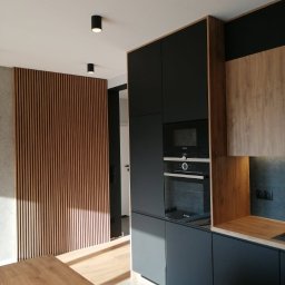 byMGdesigns - Nowoczesna kuchnia z ciemnymi, matowymi szafkami, drewnianymi akcentami i dekoracyjną ścianką z drewnianych listew, oświetlona punktowymi lampami sufitowymi.