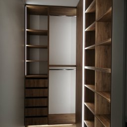 byMGdesigns - Wnętrze garderoby z drewnianymi półkami i szufladami, oświetlonymi paskami LED, widok z perspektywy wejścia.