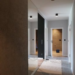 byMGdesigns - Korytarz w trakcie wykańczania z lustrem na ścianie, widoczne zabezpieczone podłogi i otwarte drzwi do łazienki z niebieskim wiadrem.