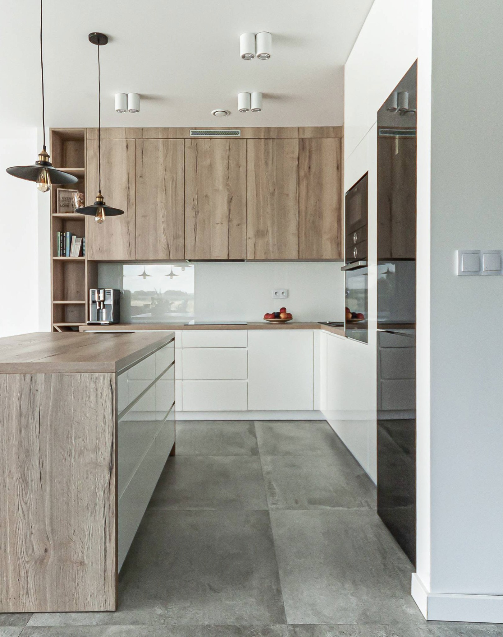 Nowoczesna kuchnia z wyspą, drewniane szafki górne i białe dolne, szare płytki podłogowe, lampy wiszące z żarówkami Edisona, ekspres do kawy na blacie, minimalistyczny design.