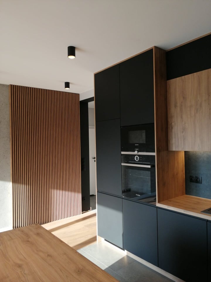 Nowoczesna kuchnia z ciemnymi, matowymi szafkami, drewnianymi akcentami i dekoracyjną ścianką z drewnianych listew, oświetlona punktowymi lampami sufitowymi.