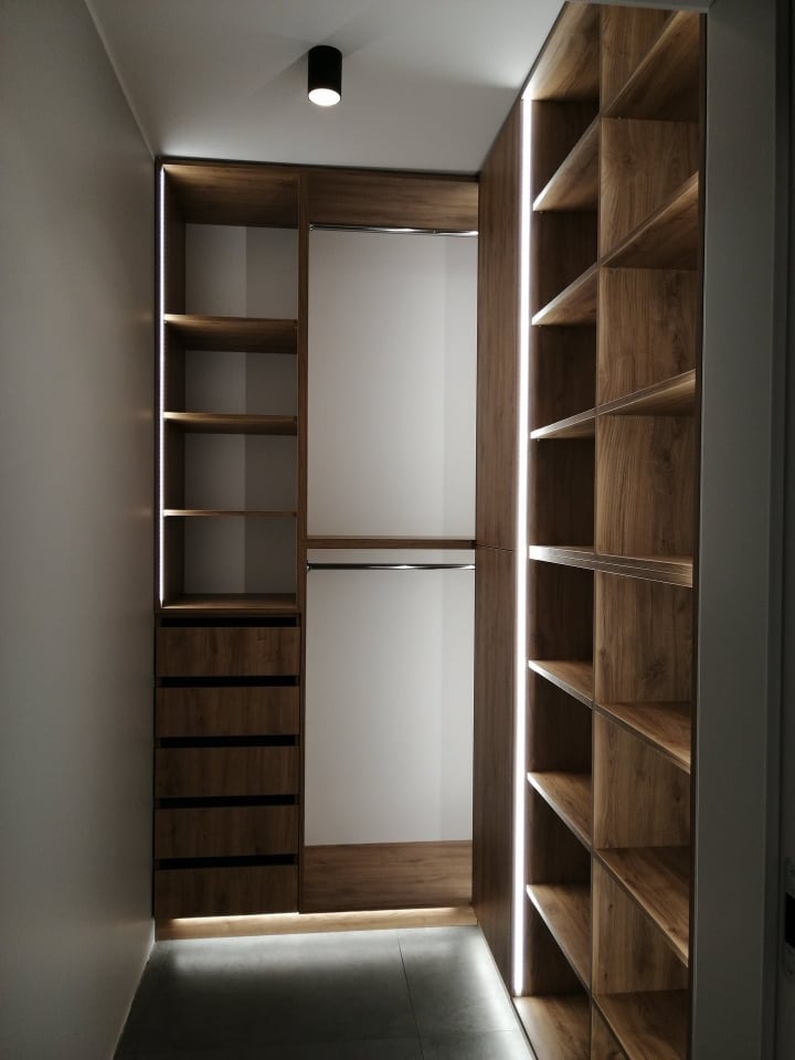 Wnętrze garderoby z drewnianymi półkami i szufladami, oświetlonymi paskami LED, widok z perspektywy wejścia.