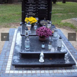 Czarny, polerowany nagrobek z wygrawerowanymi imionami i datami, ozdobiony żółtymi i fioletowymi kwiatami w doniczkach oraz zapalonymi zniczami, otoczony kostką brukową.