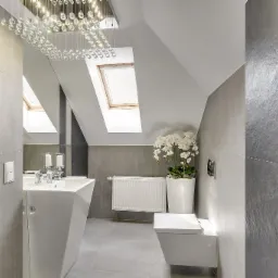 Nowoczesna łazienka na poddaszu z szarymi płytkami, wiszącą toaletą, grzejnikiem żeberkowym, umywalką na postumencie, kryształowym żyrandolem i storczykiem w doniczce.