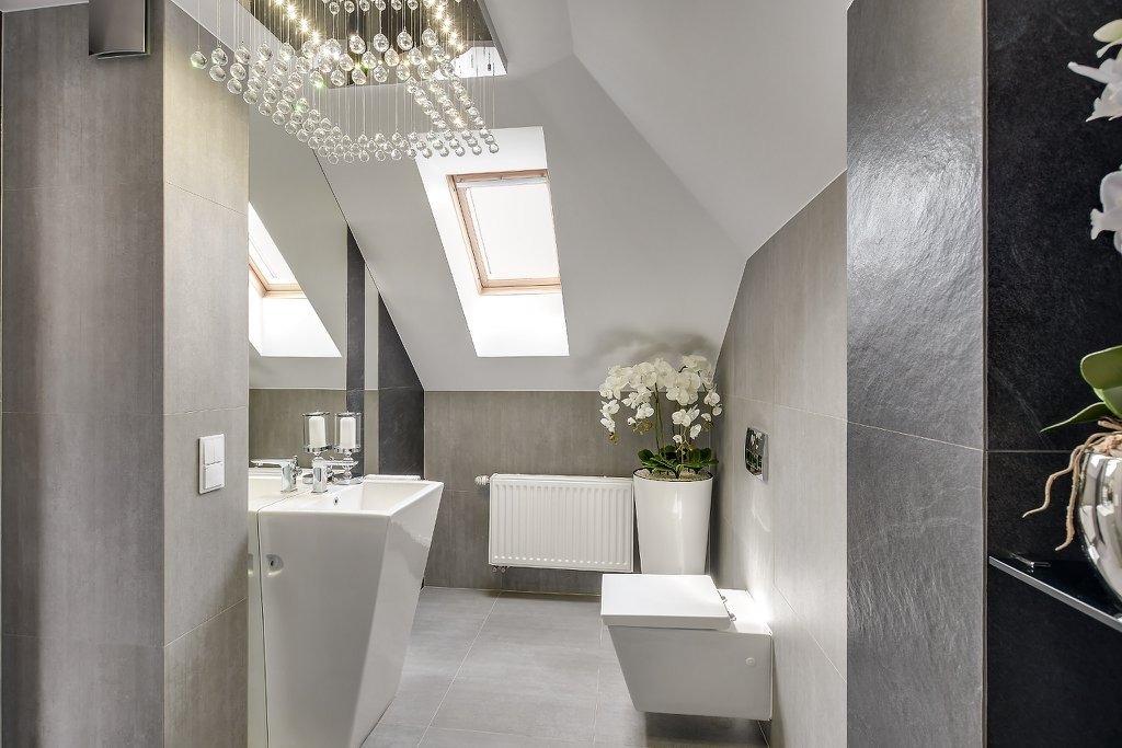 Nowoczesna łazienka na poddaszu z szarymi płytkami, wiszącą toaletą, grzejnikiem żeberkowym, umywalką na postumencie, kryształowym żyrandolem i storczykiem w doniczce.