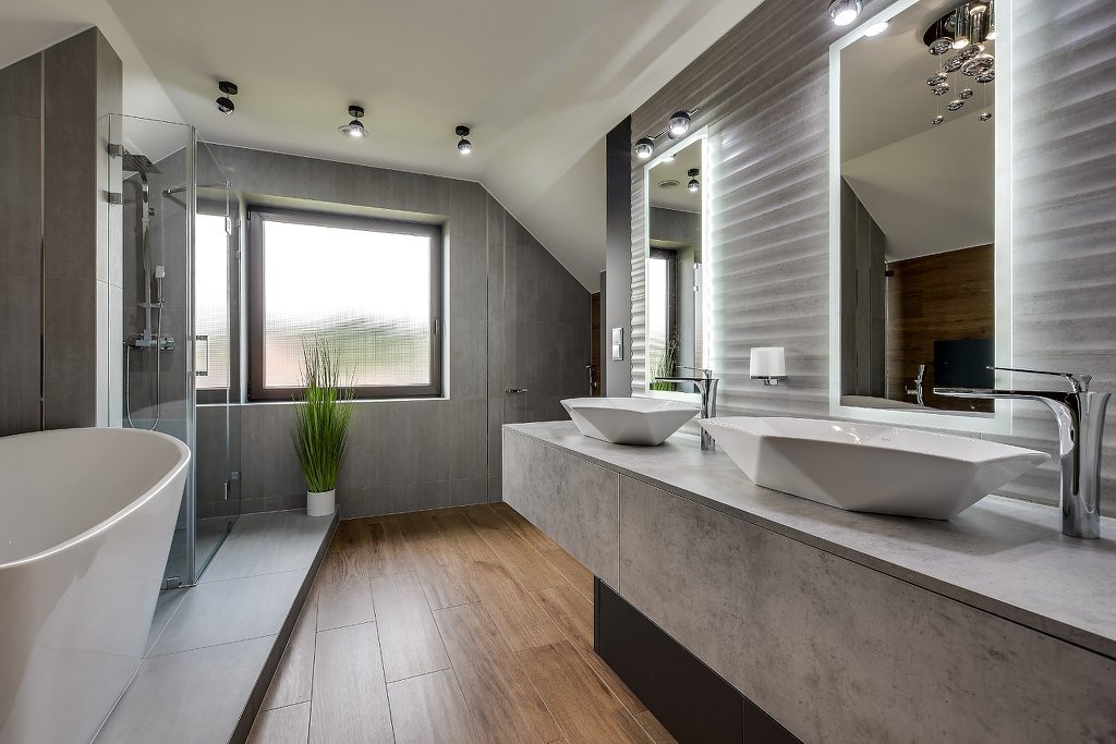 Nowoczesna łazienka na poddaszu z drewnianą podłogą, szarą ceramiką, owalną wanną, kabiną prysznicową, dwoma geometrycznymi umywalkami nablatowymi i podświetlanymi lustrami.