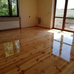 Jasna, drewniana podłoga po renowacji, z widocznym połyskiem lakieru i odbiciami światła z okien, w przestronnym pokoju z grzejnikiem i wyjściem na balkon.