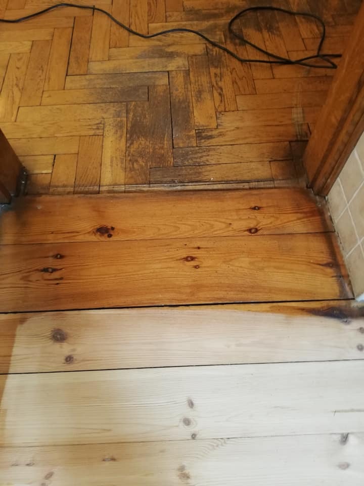 Fragment podłogi z parkietu ułożonego w jodełkę, częściowo odnowionego, z widoczną różnicą w kolorze drewna po zabiegu renowacyjnym, wraz z fragmentem progu i kablem elektrycznym.