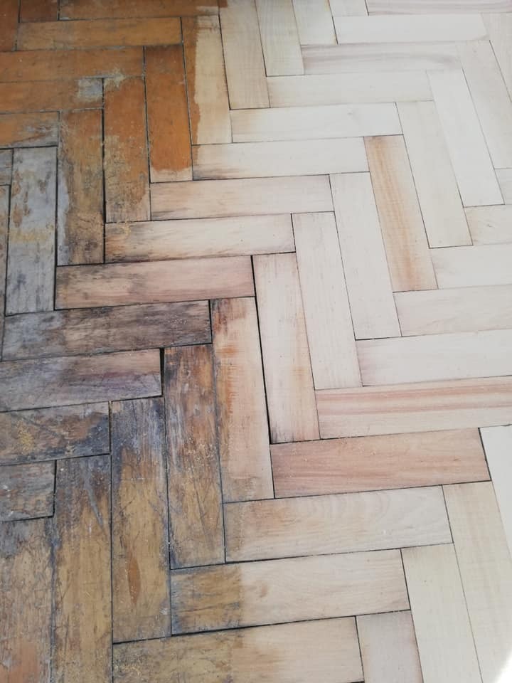Drewniana podłoga parkietowa ułożona w jodełkę, w połowie odnowiona, ukazująca różnicę między stanem przed i po renowacji, z widocznymi śladami zużycia i świeżo wyszlifowaną powierzchnią.