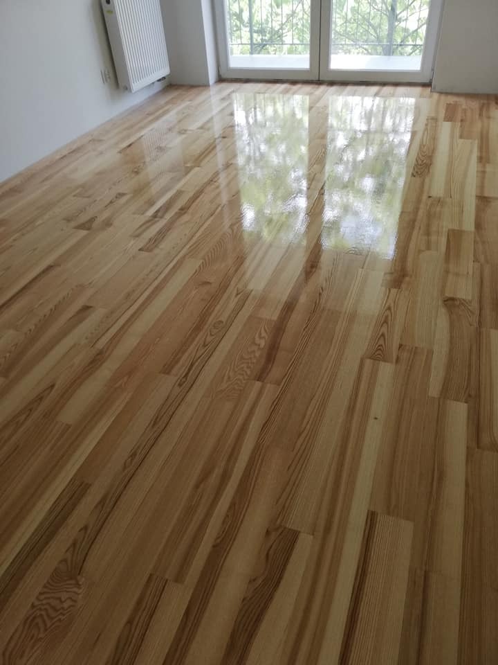 Błyszcząca, świeżo wykończona podłoga z jasnego drewna, ułożona wzdłuż okna z widocznymi refleksami światła, grzejnik na ścianie w tle.