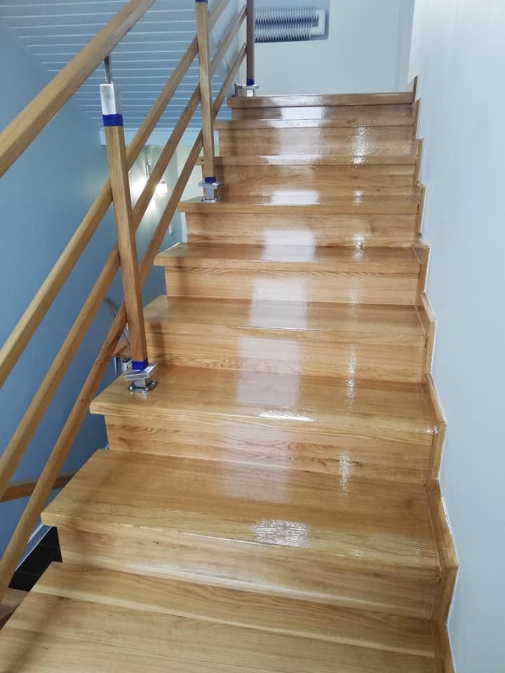 Błyszczące, drewniane schody po renowacji, z jasną balustradą i niebieskimi akcentami, widoczne ślady po cyklinowaniu na kilku stopniach.