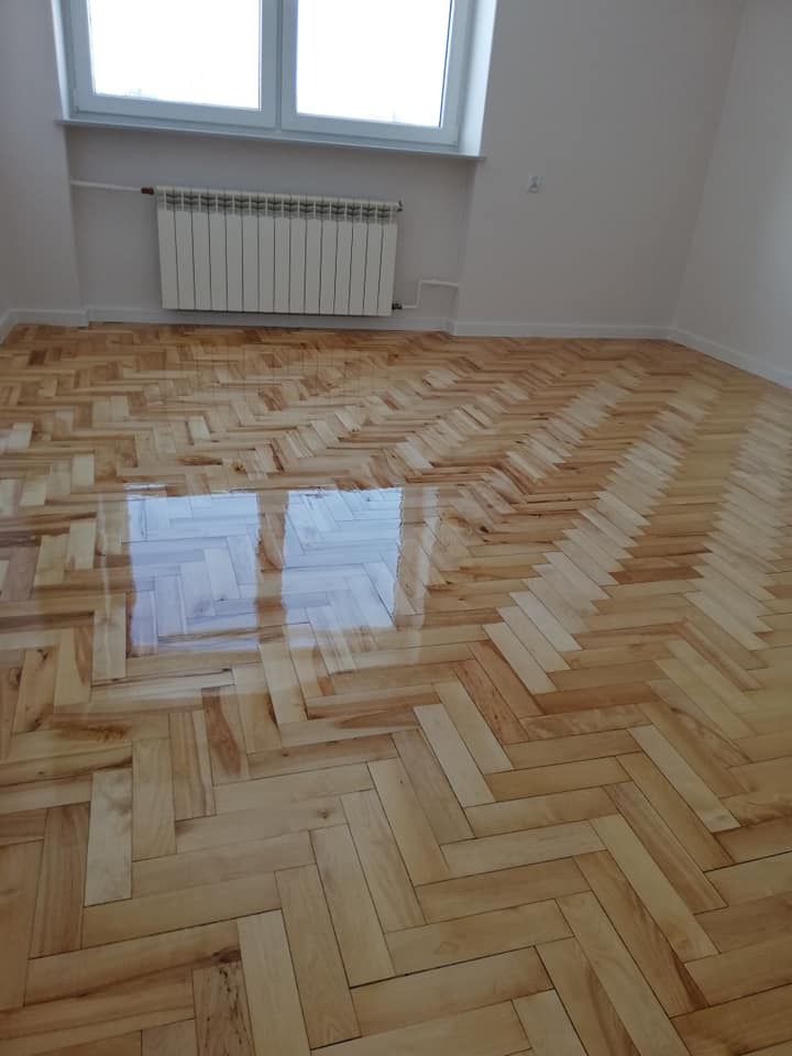 Świeżo polakierowany parkiet ułożony w jodełkę klasyczną, odbijający światło okna w pustym, odnowionym pokoju z białymi ścianami i grzejnikiem.