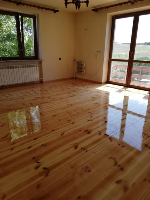 Jasna, drewniana podłoga po renowacji, z widocznym połyskiem lakieru i odbiciami światła z okien, w przestronnym pokoju z grzejnikiem i wyjściem na balkon.
