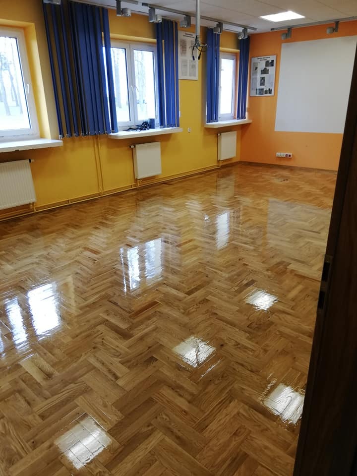 Błyszcząca, odnowiona podłoga parkietowa ułożona we wzór jodełki w jasnym pomieszczeniu z żółtymi ścianami i niebieskimi zasłonami.