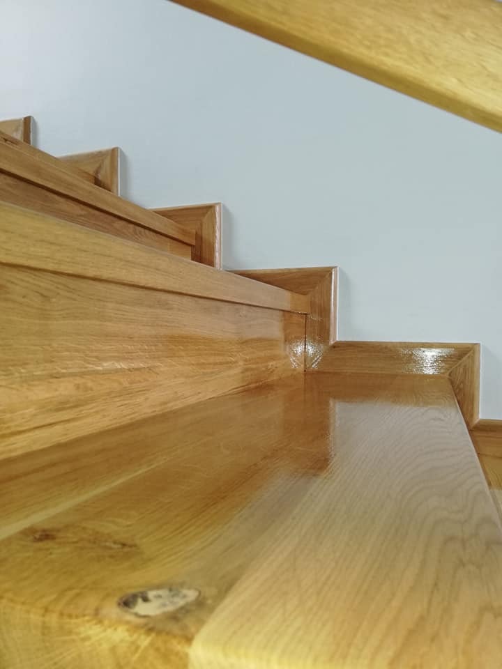 Błyszczące, drewniane schody po renowacji, widoczne detale usłojenia drewna i świeżo nałożony lakier, minimalistyczne białe tło ściany.