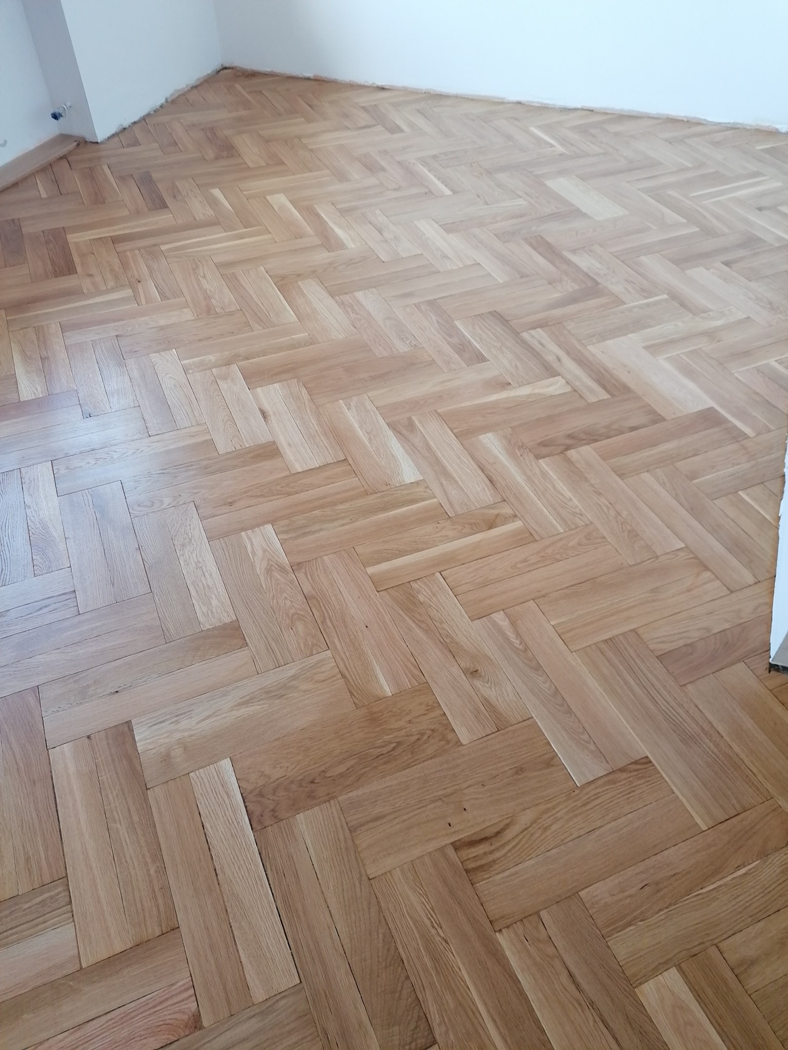 Jasny, dębowy parkiet ułożony w jodełkę francuską, świeżo po lakierowaniu, widoczny fragment białej ściany w rogu pomieszczenia.