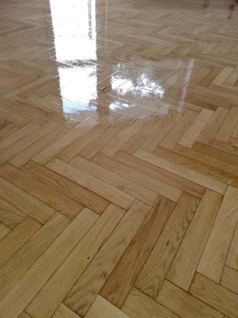 Świeżo polakierowany parkiet ułożony w jodełkę, odbijający światło okna, ukazujący naturalne usłojenie drewna.