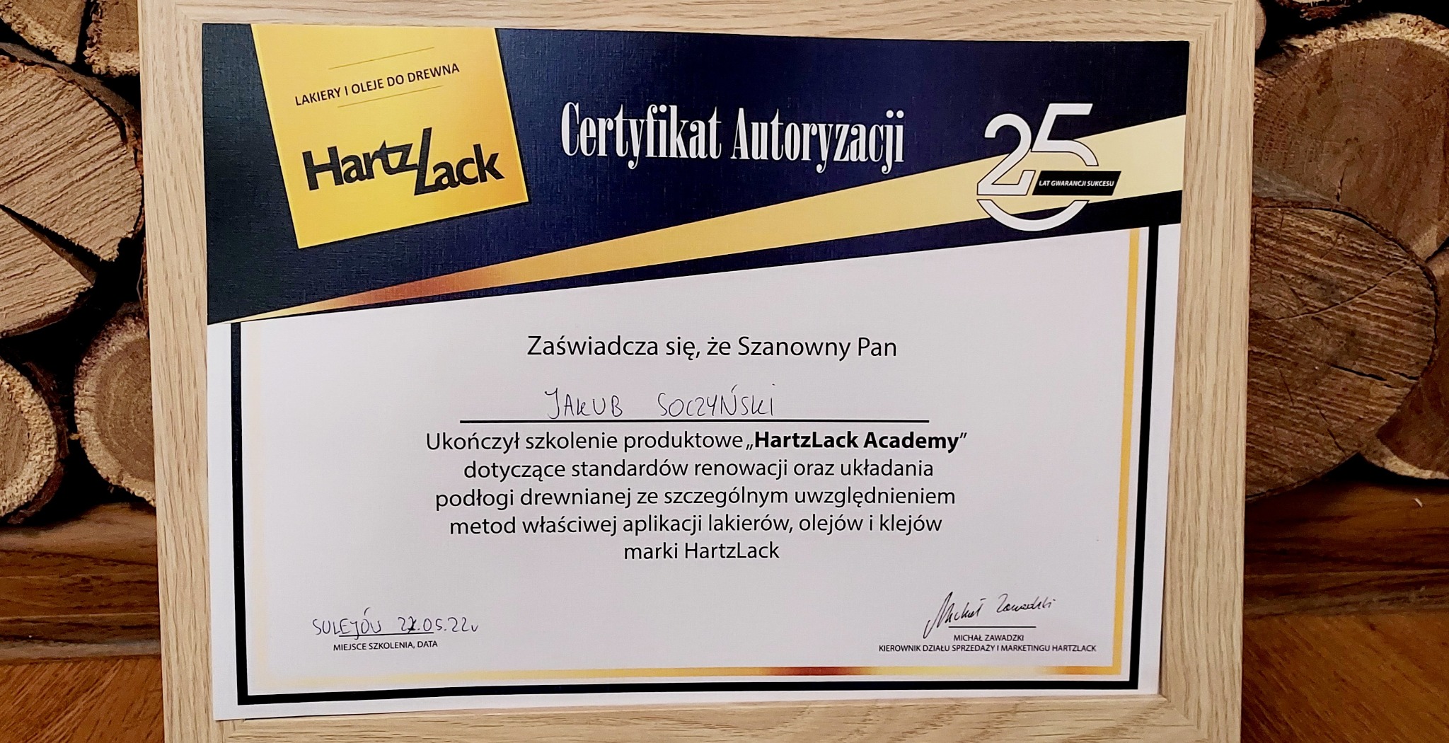 Oprawiony certyfikat autoryzacji HartzLack na tle ułożonych kawałków drewna, potwierdzający ukończenie szkolenia z zakresu renowacji i układania podłóg drewnianych z użyciem lakierów, olejów...