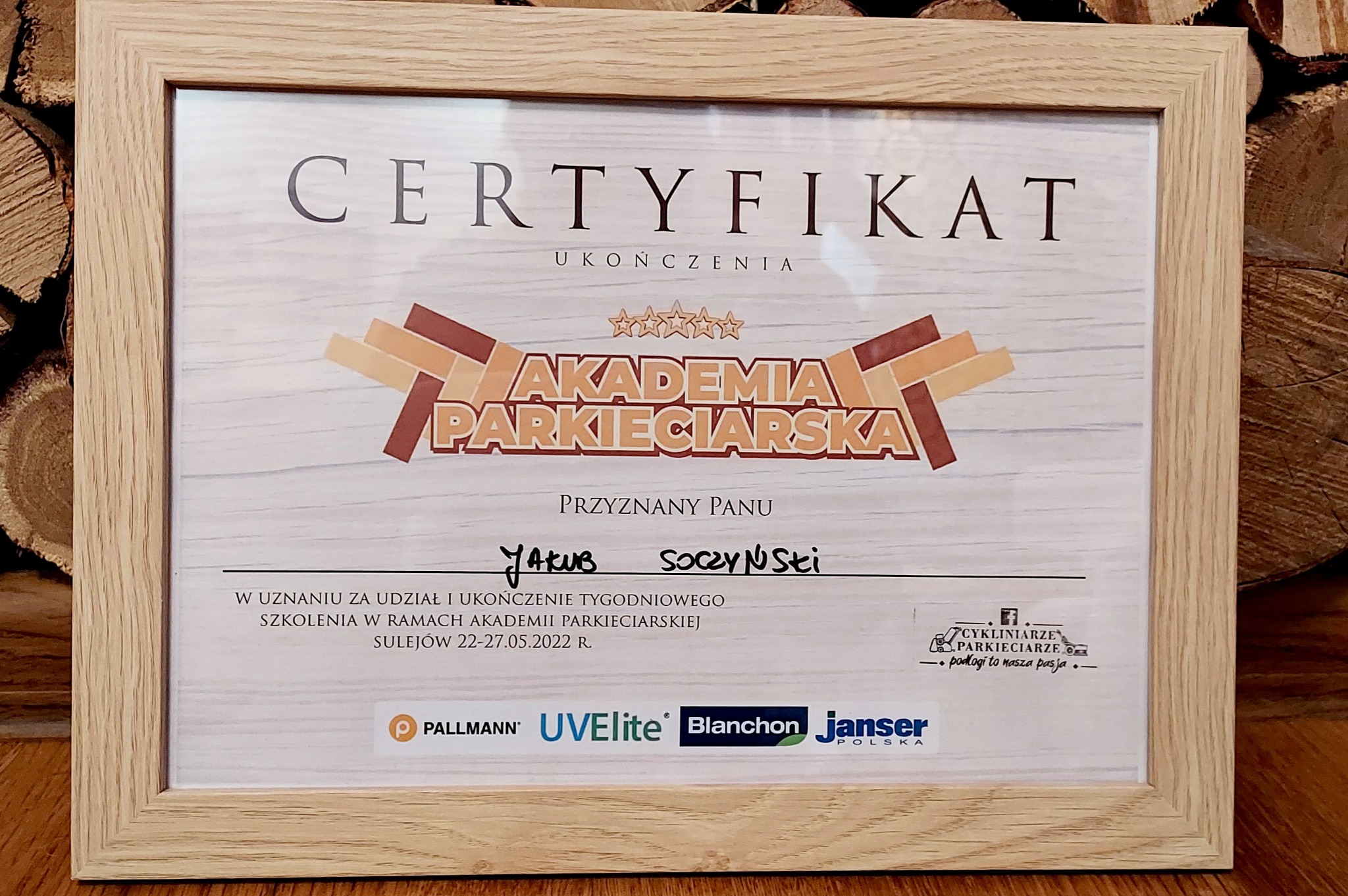 Oprawiony w drewnianą ramkę certyfikat ukończenia Akademii Parkieciarskiej, przyznany Panu Jakubowi Soczyńskiemu, z logo firmy Cykliniarze Parkieciarze i sponsorów szkolenia, na tle stosu drewna.