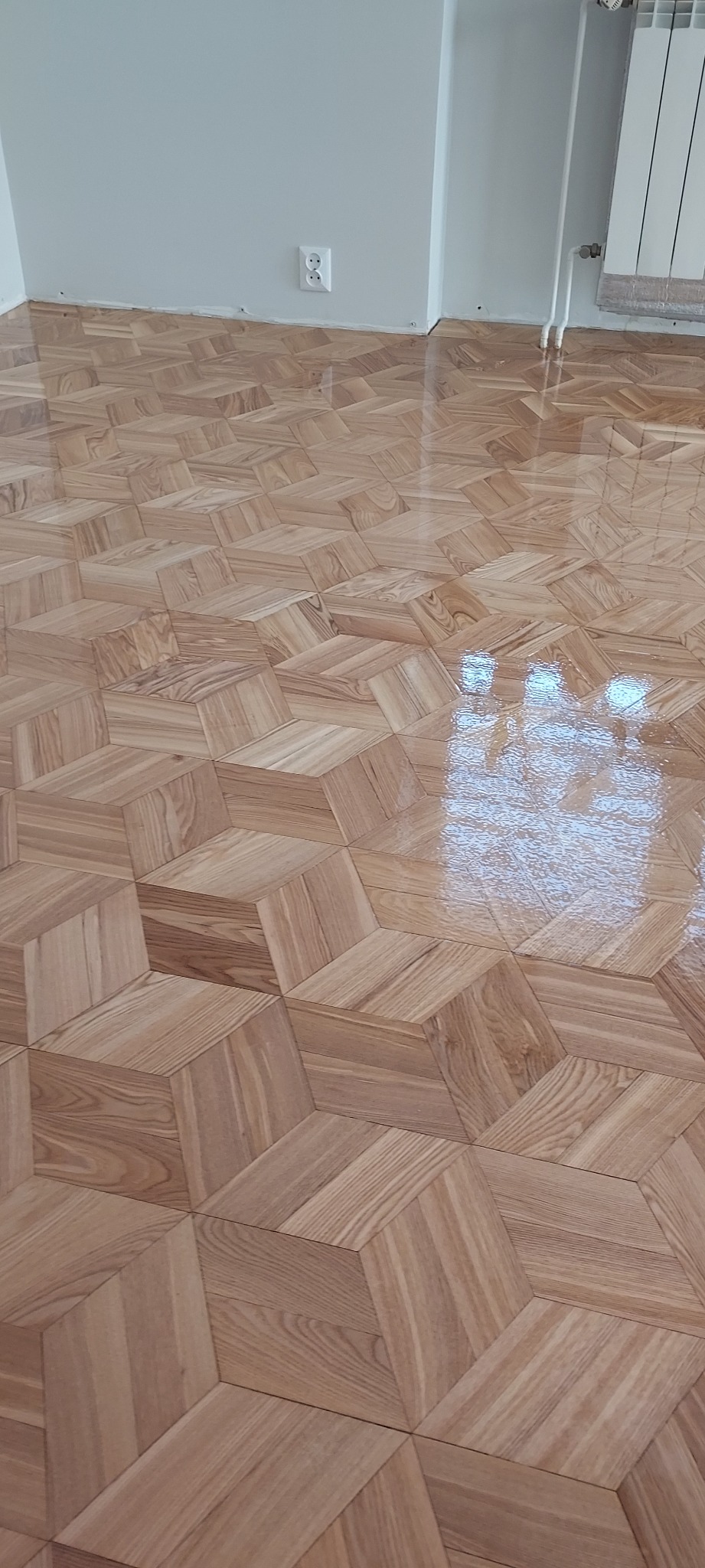 Ułożony parkiet w geometryczny wzór 3D, z widocznym połyskiem lakieru, jasne ściany i grzejnik w tle.