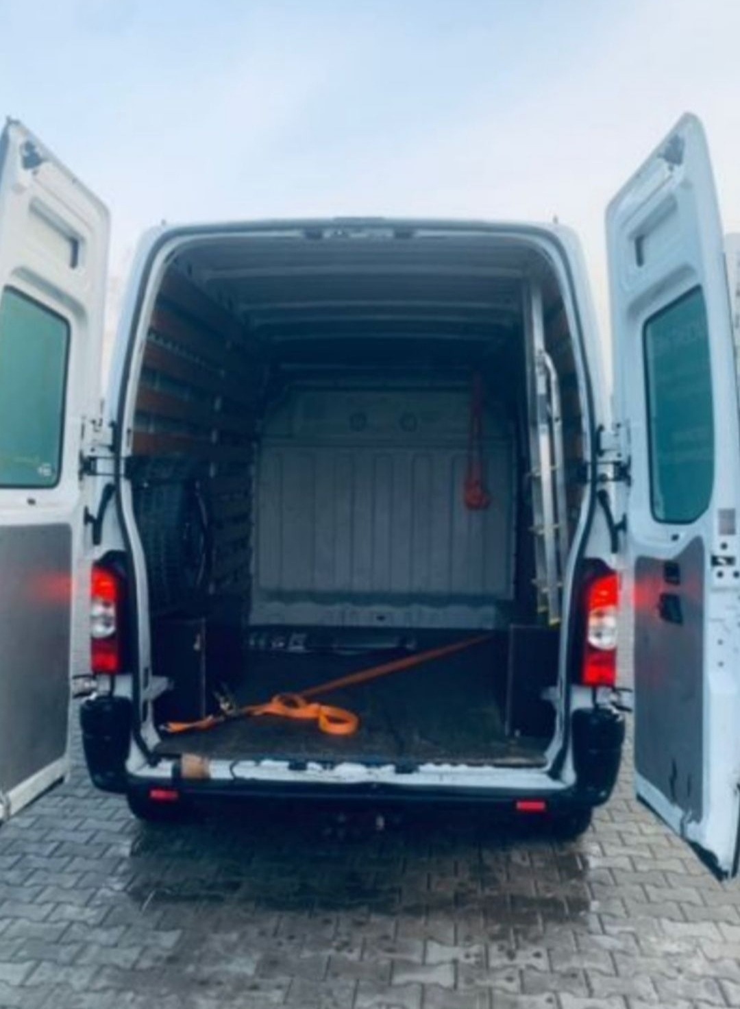 Wnętrze białego busa z otwartymi drzwiami, widoczne pasy transportowe, drabina i zapasowe koło