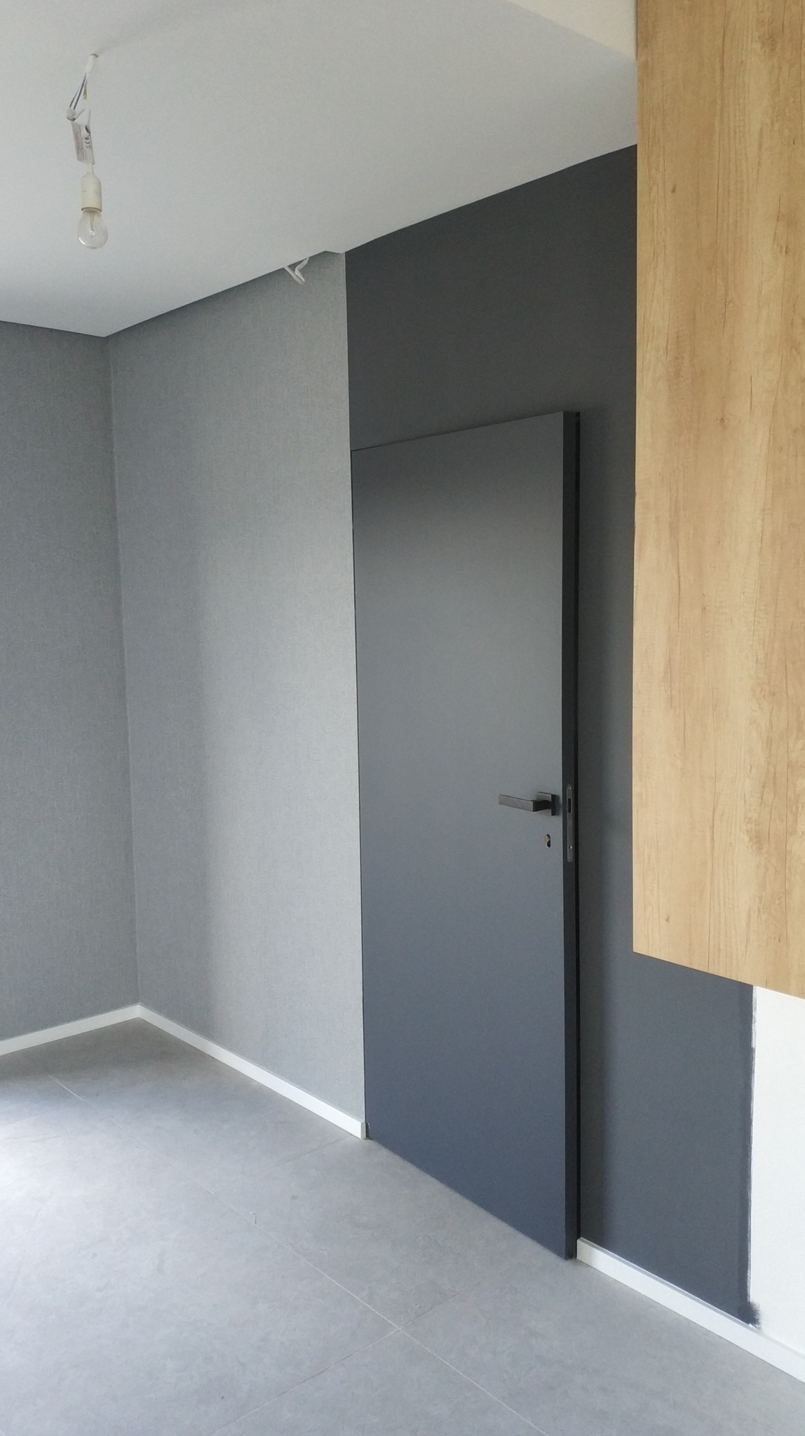 Wnętrze pokoju po remoncie: szara ściana z tapetą strukturalną, ciemnoszara ściana z drzwiami wewnętrznymi, fragment drewnianej okładziny ściennej, szara podłoga z płytek, białe listwy przypodłogowe.
