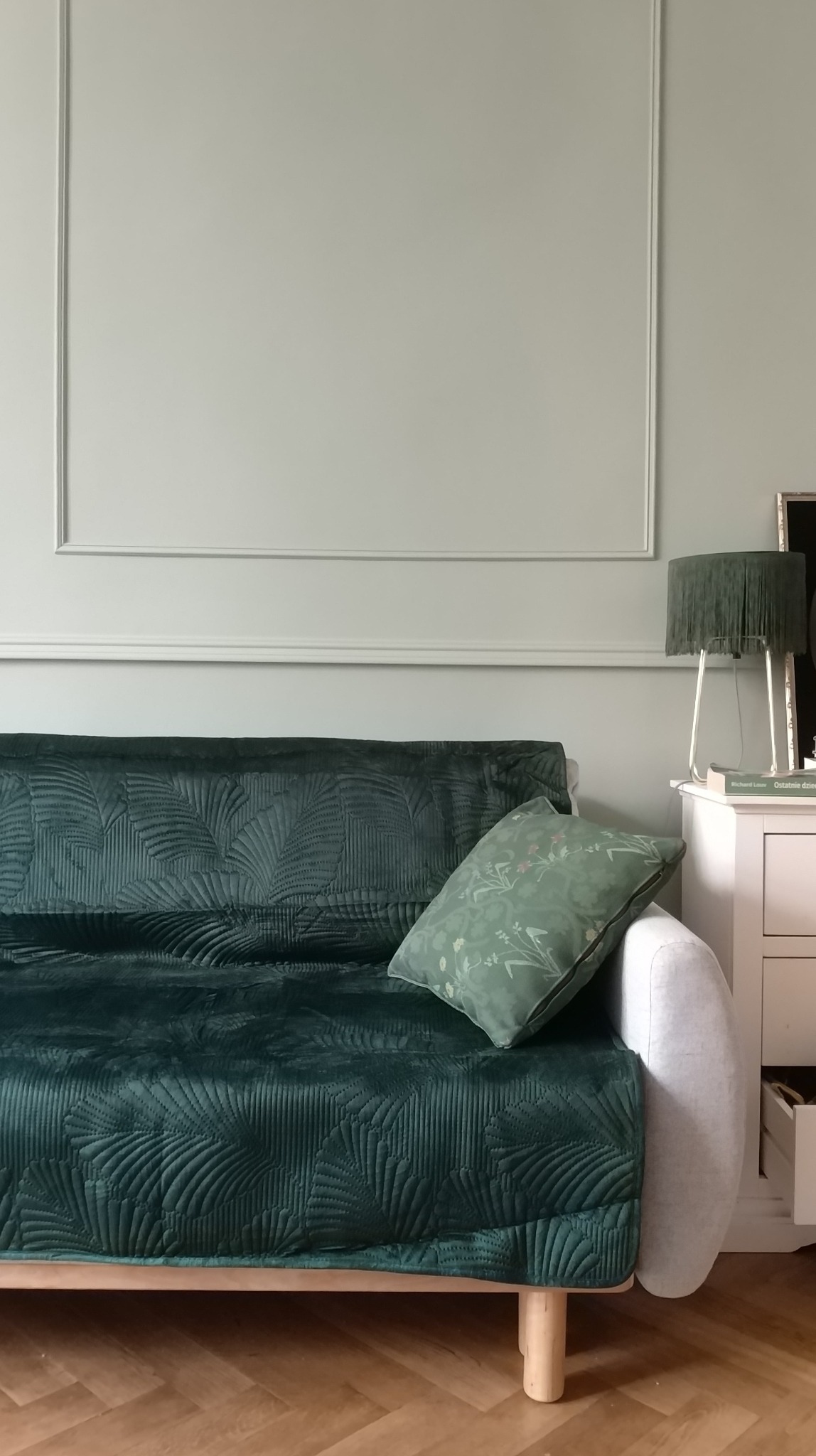 Stylowe wnętrze z zieloną sofą o geometrycznym wzorze, poduszką w kwiaty i lampką z frędzlami na białej komodzie, na tle jasnej ściany z dekoracyjną listwą.