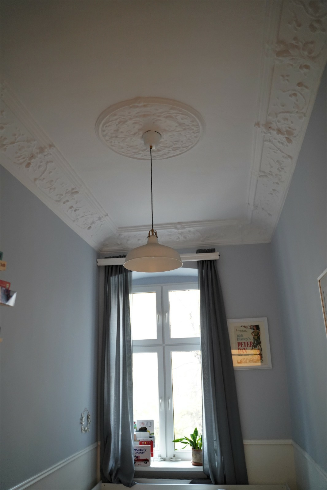 Pokój z szarą ścianą, białym sufitem z dekoracyjną sztukaterią i centralnie umieszczoną okrągłą lampą wiszącą, okno z szarymi zasłonami.