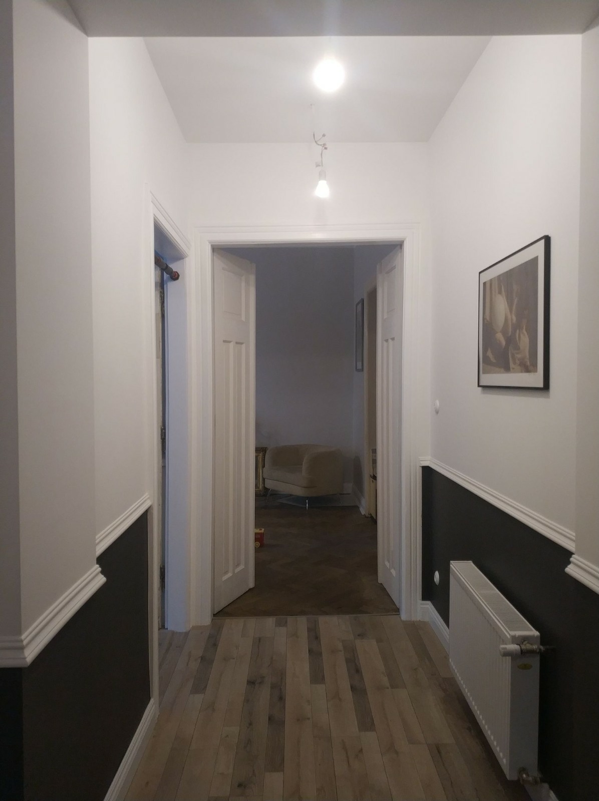 Korytarz po odświeżeniu: ściany pomalowane na biało z dolną częścią w kolorze ciemnego grafitu, drewniana podłoga, białe drzwi, grzejnik i obraz na ścianie.