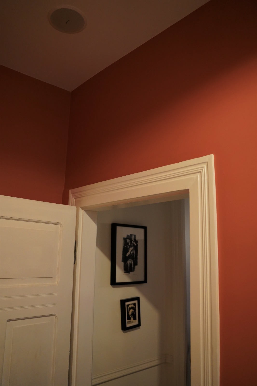 Pomarańczowa ściana z białymi drzwiami i futryną, w tle widoczne grafiki w czarnych ramkach na białej ścianie.