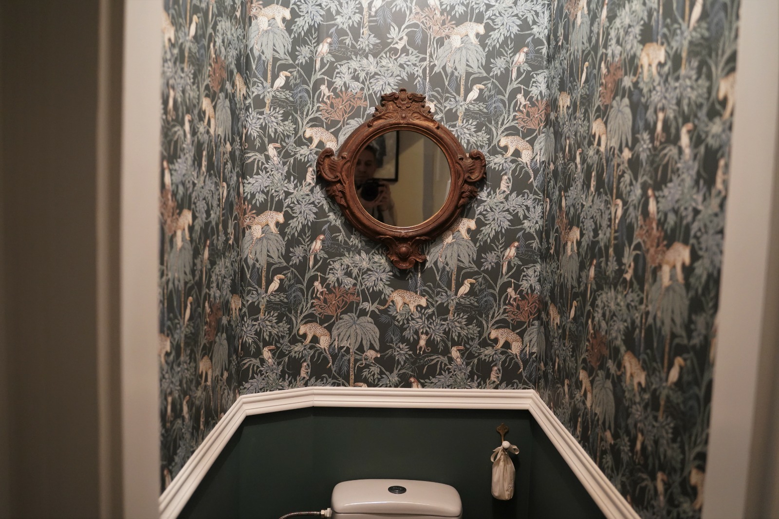 Wzorzysta tapeta z motywem dżungli w łazience, ozdobne lustro w drewnianej ramie, biała listwa przypodłogowa i fragment toalety.