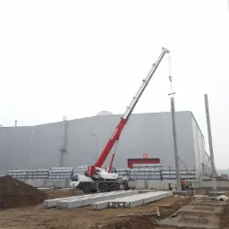 Montaż betonowego słupa konstrukcyjnego przy użyciu dźwigu samochodowego na placu budowy, widoczni pracownicy w kaskach i kamizelkach odblaskowych, w tle hala magazynowa.