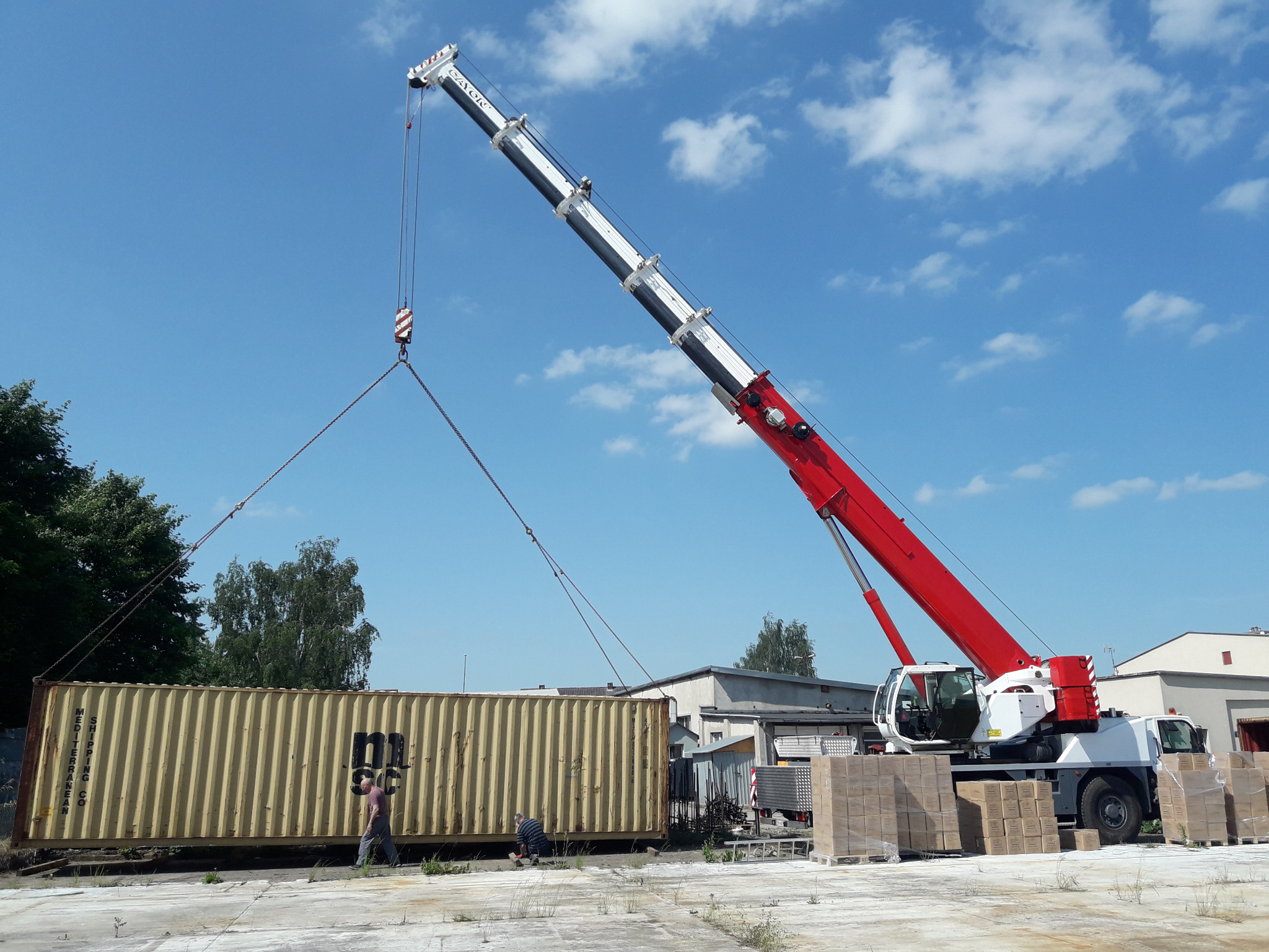 Żółty kontener transportowy podnoszony przez czerwono-biały dźwig samochodowy na tle błękitnego nieba z chmurami, w pobliżu stoją mężczyźni i palety z kartonami.