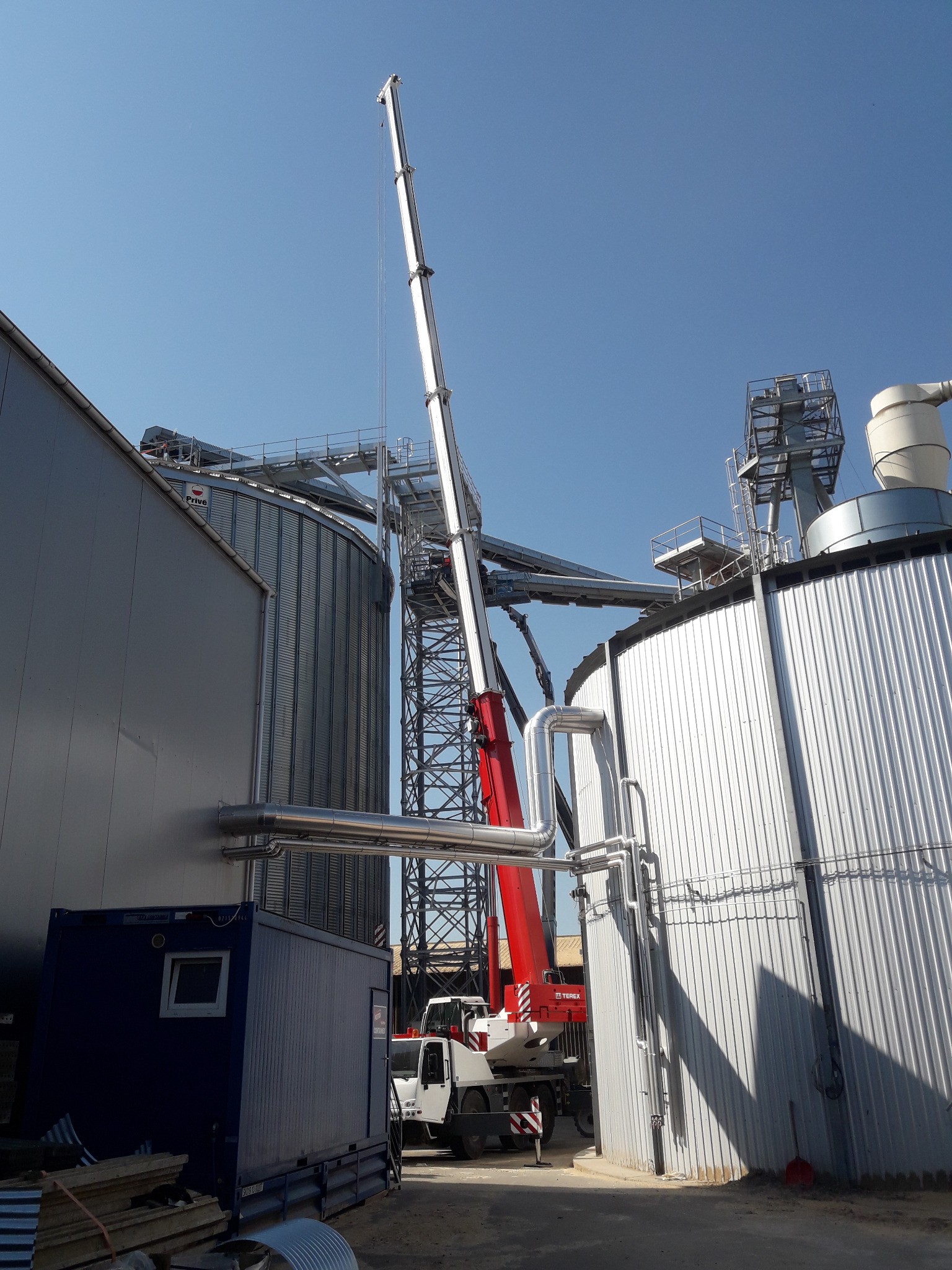 Montaż silosów zbożowych z użyciem dźwigu Terex, widoczne elementy konstrukcyjne i rurociągi na tle błękitnego nieba.
