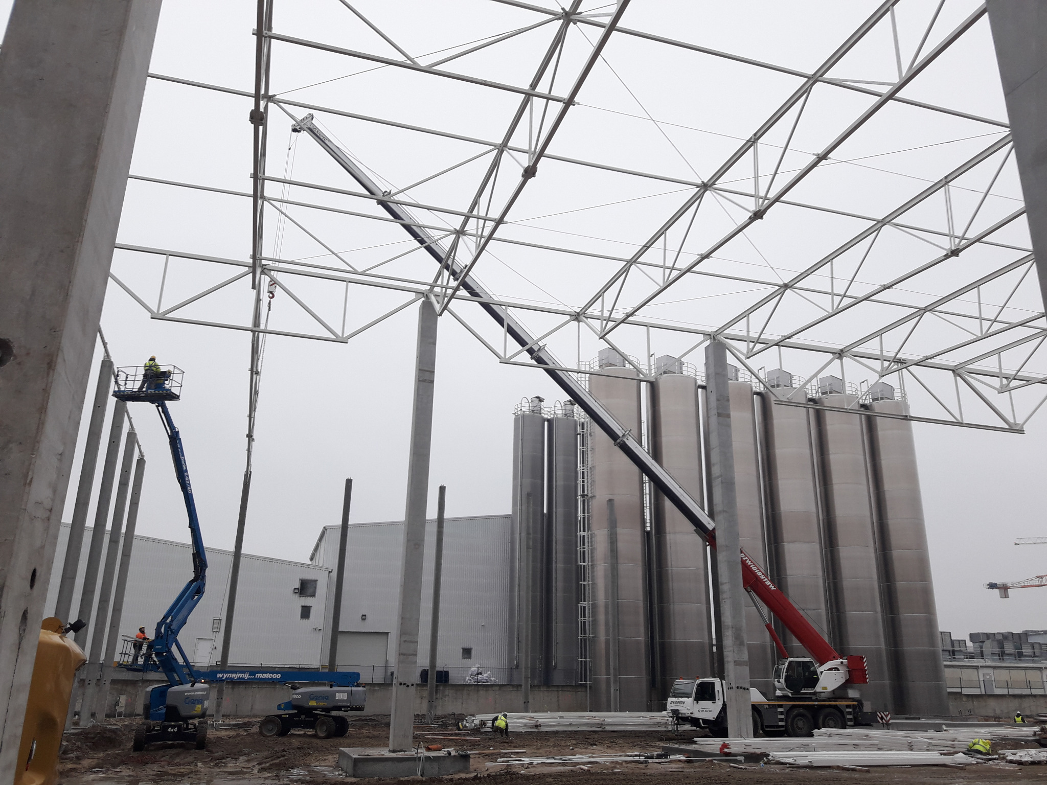 Montaż konstrukcji stalowej hali przemysłowej z użyciem dwóch podnośników koszowych i dźwigu samochodowego; widoczne słupy betonowe, elementy konstrukcji dachu i zbiorniki przemysłowe w tle.