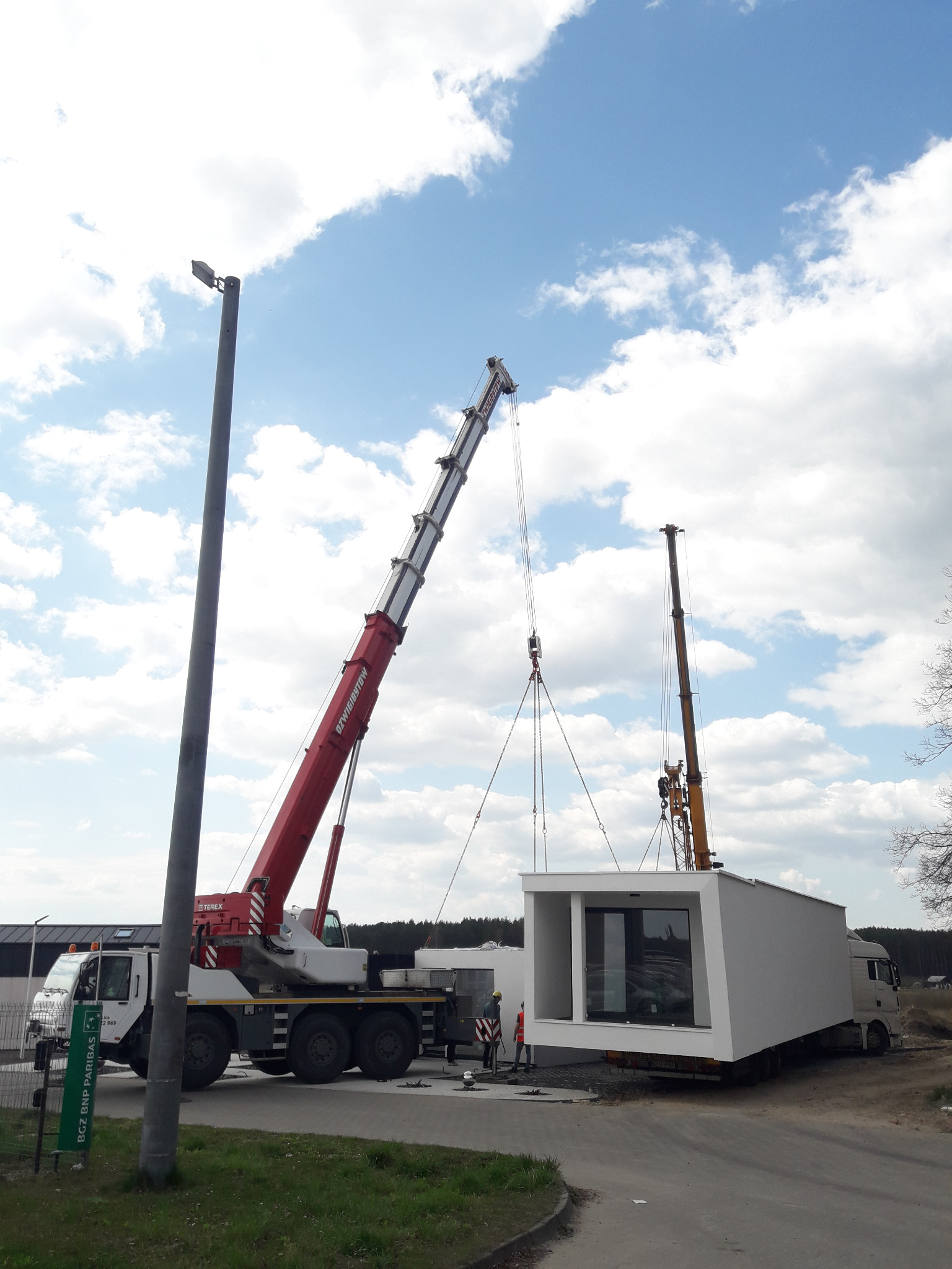 Dwa dźwigi przenoszą modułowy, biały budynek z dużymi oknami na naczepę tira; błękitne niebo z chmurami w tle.