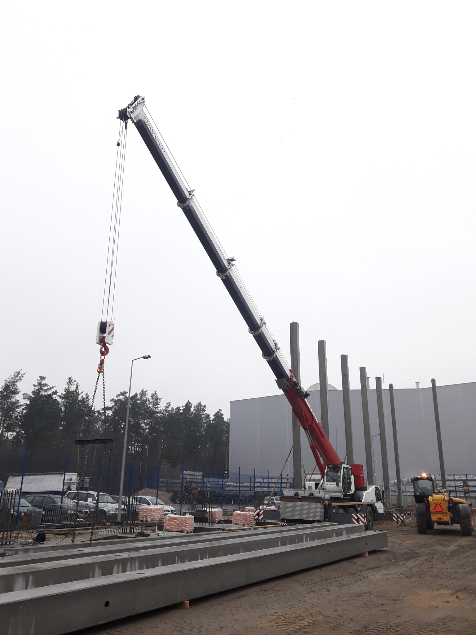 Żuraw samochodowy podnosi elementy konstrukcyjne w trakcie budowy hali, widoczny ciągnik i ułożone bloczki.