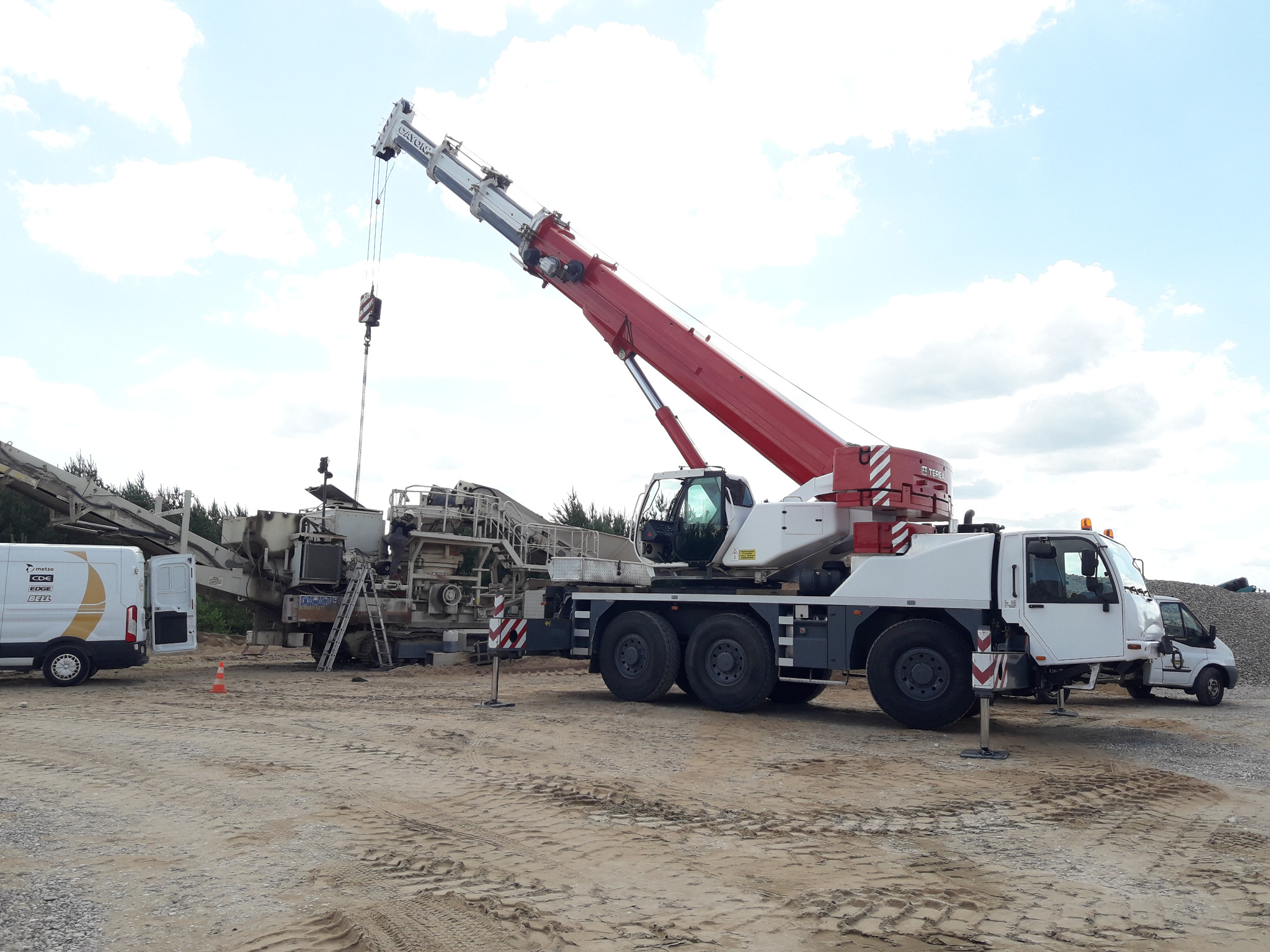 Mobilny żuraw z wysięgnikiem ustawiony obok maszyny do kruszenia kamienia, widoczne ślady opon na piaszczystym podłożu i samochód dostawczy w tle.