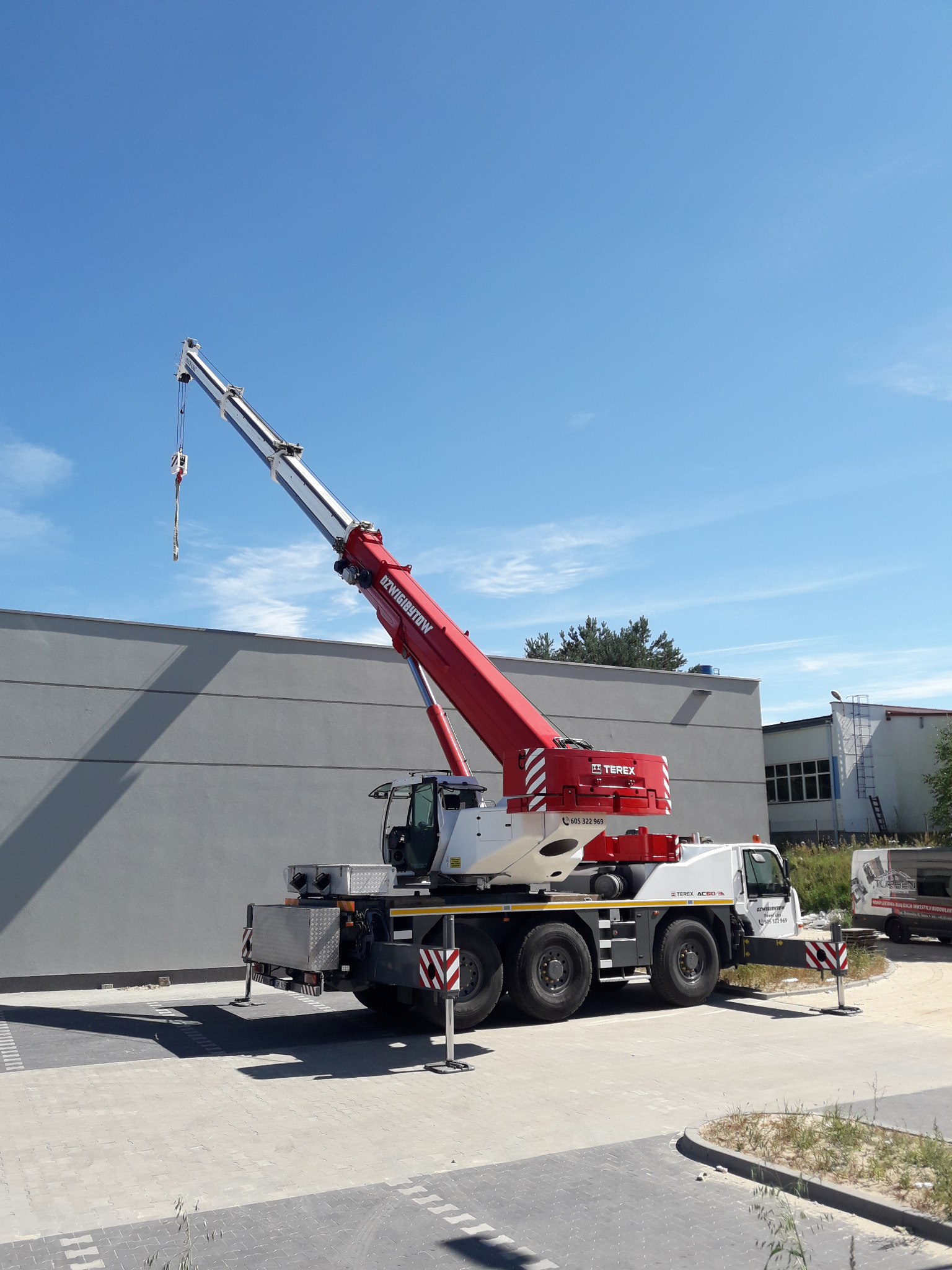 Żuraw samojezdny Terex AC 300/6 z wysuniętym teleskopowym ramieniem na tle szarego budynku przemysłowego i błękitnego nieba.