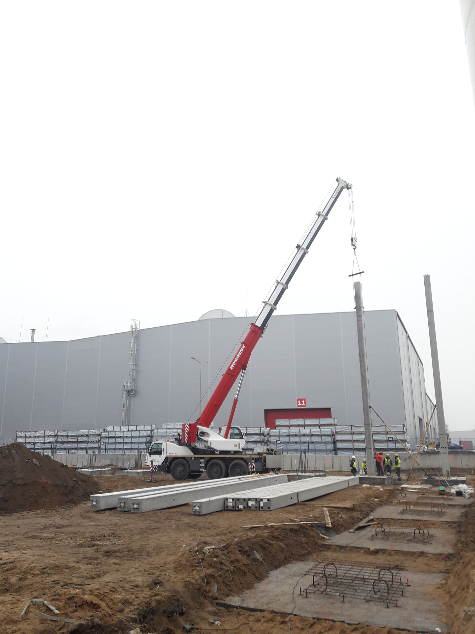 Montaż betonowego słupa konstrukcyjnego przy użyciu dźwigu samochodowego na placu budowy, widoczni pracownicy w kaskach i kamizelkach odblaskowych, w tle hala magazynowa.