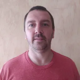 Portret mężczyzny na tle beżowej ściany z efektem dekoracyjnym, w czerwonej koszulce, z widocznym znakiem wodnym telefonu w dolnej części kadru.