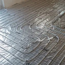 Białe rury ogrzewania podłogowego ułożone na srebrnej folii z nadrukowaną czarną siatką i logo, w surowym pomieszczeniu z oświetleniem od okna.