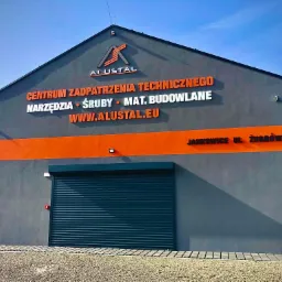 Budynek firmy Alustal z szarymi ścianami, pomarańczowym pasem i napisem informującym o asortymencie: narzędzia, śruby, materiały budowlane.