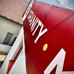 Gościu od reklamy - Czerwony szyld reklamowy z białym napisem 'IB Gravity' i żółtą kropką, zamontowany na budynku z widocznym fragmentem dachu i niebem w tle.