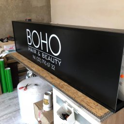 Gościu od reklamy - Czarny szyld reklamowy BOHO Hair & Beauty z białym napisem, widoczny w trakcie montażu w warsztacie, obok rolki zielonej folii i narzędzi.