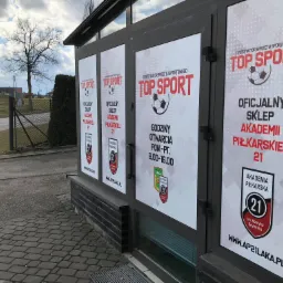 Okna lokalu oklejone plakatami informacyjnymi 'Top Sport - oficjalny sklep Akademii Piłkarskiej 21', z godzinami otwarcia, widok z zewnątrz na chodnik z kostki brukowej i fragment boiska w tle.