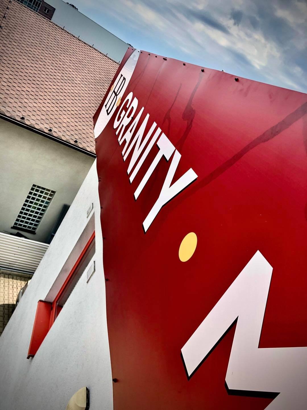 Czerwony szyld reklamowy z białym napisem 'IB Gravity' i żółtą kropką, zamontowany na budynku z widocznym fragmentem dachu i niebem w tle.