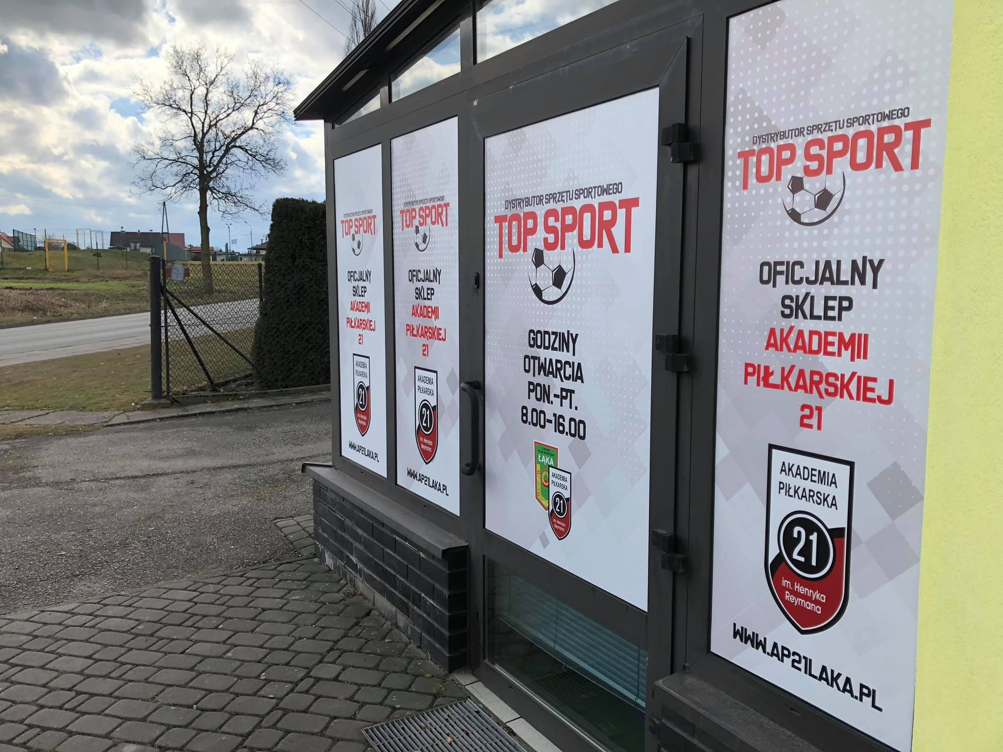 Okna lokalu oklejone plakatami informacyjnymi 'Top Sport - oficjalny sklep Akademii Piłkarskiej 21', z godzinami otwarcia, widok z zewnątrz na chodnik z kostki brukowej i fragment boiska w tle.